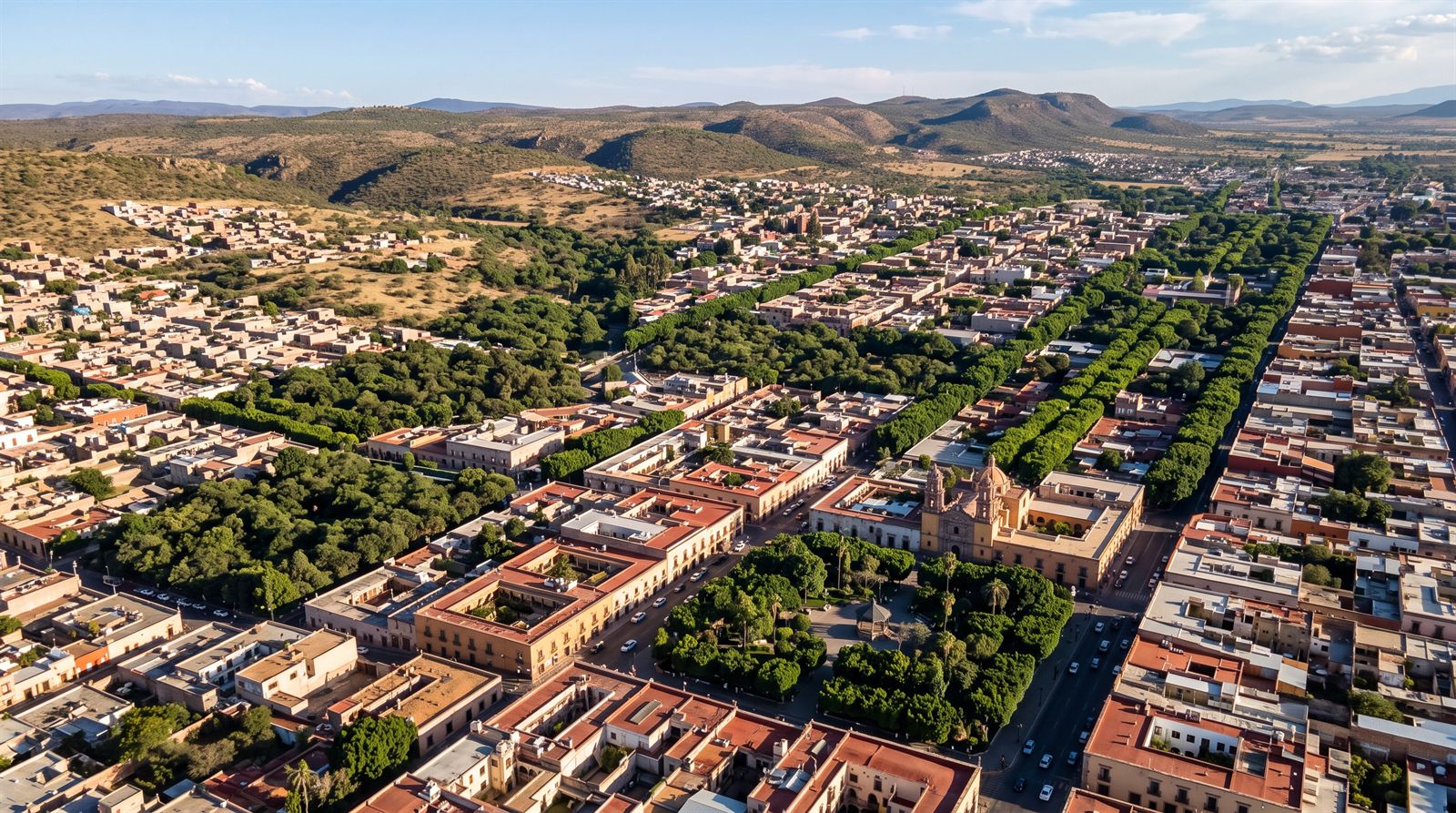 Vista aérea de una ciudad colonial con calles arboladas que forman una red verde continua entre las manzanas, con cerros al fondo